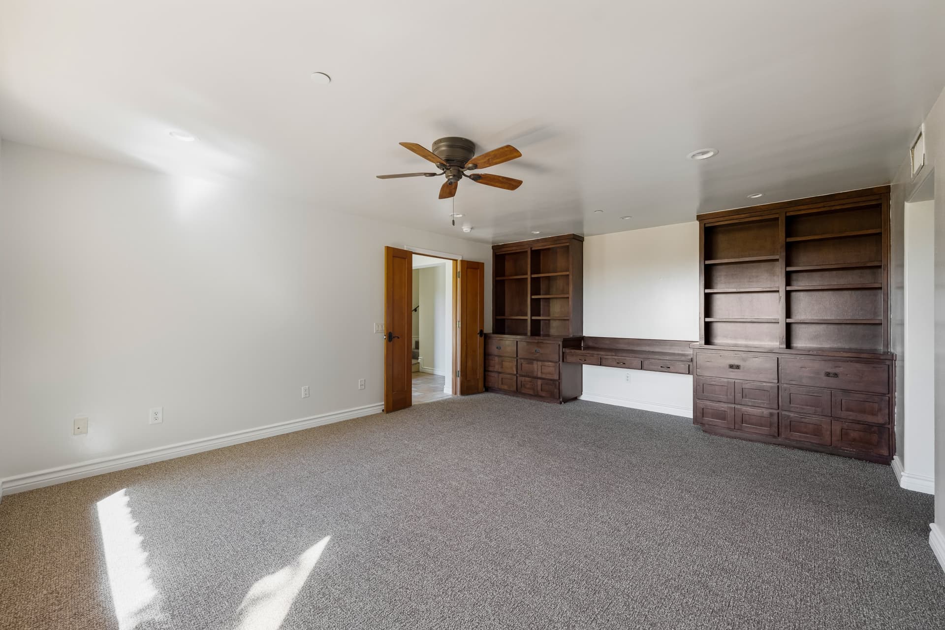Den with built-in shelving, a ceiling fan and sun patches on the carpet