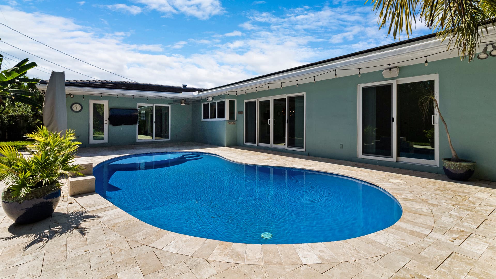 Backyard pool in daylight with unlit string lights along the roofline